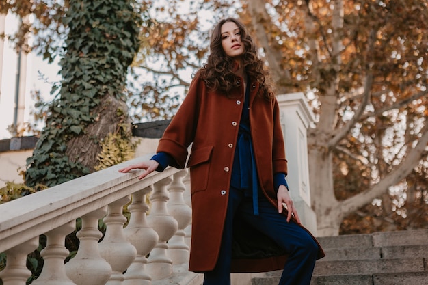 Free photo beautiful stylish smiling skinny woman with curly hair walking in street stairs dressed in warm brown coat and blue suit, autumn trendy fashion street style