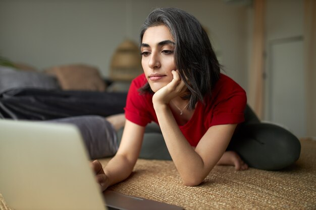 Beautiful stylsih young female with grayish hair and facial piercing sitting on carpet in front of open laptop computer, surfing internet using wireless connection, watching yoga tutorial online.