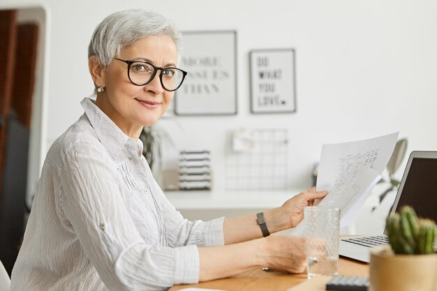 Beautiful successful confident mature businesswoman with short gray hair working at her office, using portable computer holding papers in her hands, studying financial report, smiling 