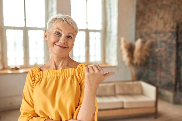 Beautiful successful middle aged female real estate agent with pixie haircut making thumbs up gesture, pointing finger at cozy living room with stylish interior design, offering apartment for sale