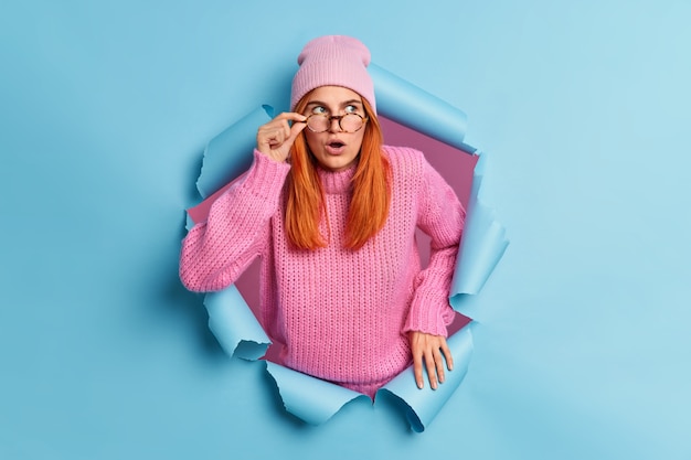 Beautiful surprised redhead young woman keeps mouth opened sees something unbelievable amazes by unexpected offer wears pink hat and sweater breaks through blue paper