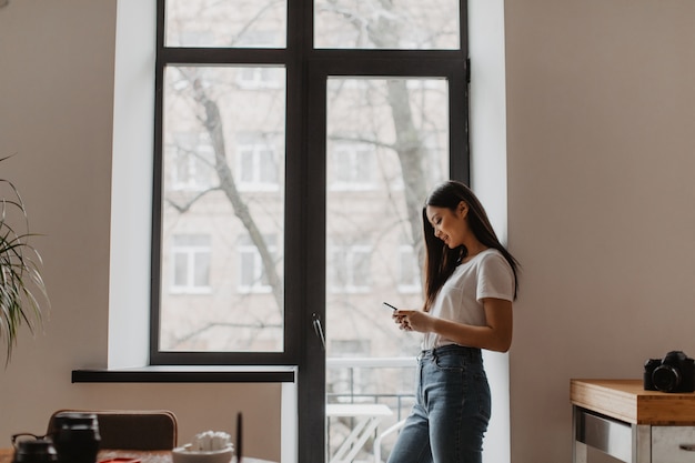 Free Photo beautiful tanned woman chats in smartphone, leaning on white wall by window