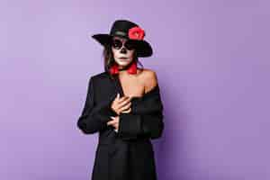 Free photo beautiful tanned woman with face art dressed in black jacket and hat, posing on lilac wall.