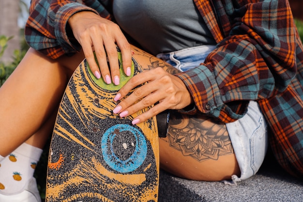 Free photo beautiful tattooed fit woman in jean shorts, plaid shirt sit on stairs at sunset light holding longboard