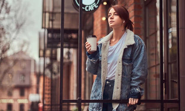 A beautiful tattooed girl wearing a denim coat holding cup with takeaway coffee outside near the cafe.