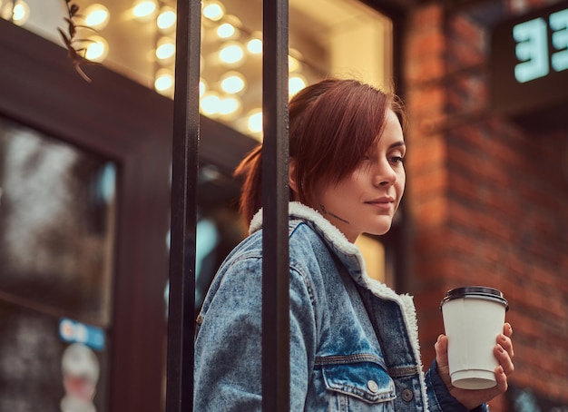 Free photo a beautiful tattooed girl wearing a denim coat holding cup with takeaway coffee outside near the cafe.