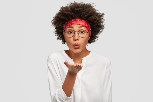 Beautiful tender black woman with Afro haircut, blows airkiss, keeps palm towards mouth, expresses love on distance