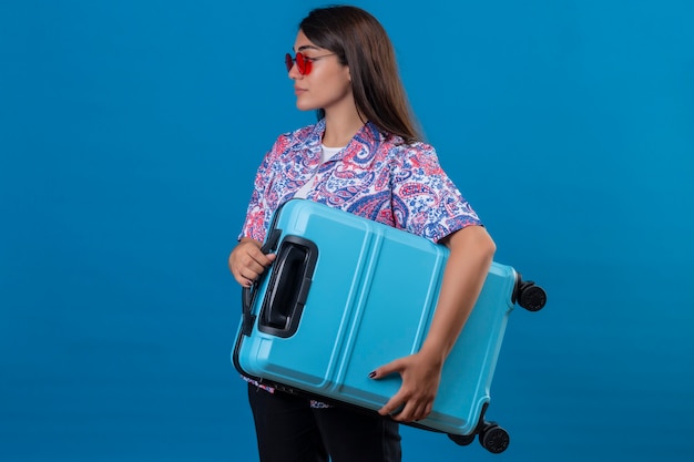 Free Photo beautiful tourist woman wearing red sunglasses holding travel suitcase looking confident standing sideways over isolated blue space