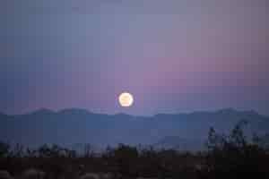 Free photo beautiful view of a full moon in the evening above the silhouettes of the mountains and greenery