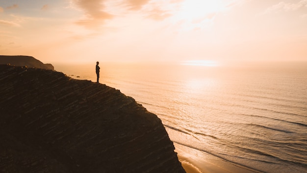 Free photo beautiful view of a person standing on a cliff over the ocean at sunset in algarve, portugal