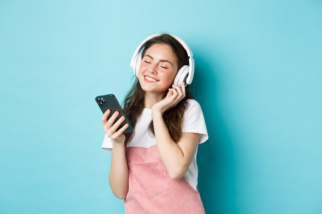 Free photo beautiful woman enjoying song in headphones, close eyes and smile while listening music in headphones, holding smartphone in hand, standing over blue background.