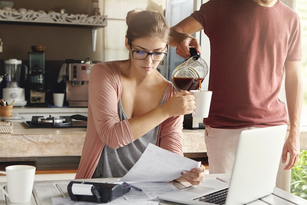 Free Photo beautiful woman in glasses holding piece of paper, doing paperwork and paying taxes at kitchen table with laptop pc and calculator on it. her husband standing beside her and adding coffee in her mug