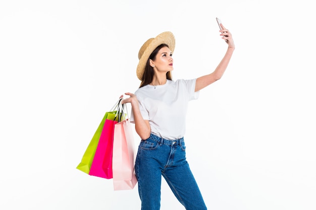 Beautiful woman holding shopping bags and taking selfie with cell phone isolated on white wall