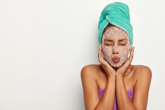 Free photo beautiful woman keeps lips folded and eyes closed, wears towel on head, makes mask for peeling face after taking shower, has beauty treatments, models over white wall, free space