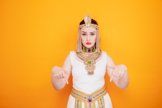 Beautiful woman like cleopatra in ancient egyptian costume confused pointing with index fingers down on orange