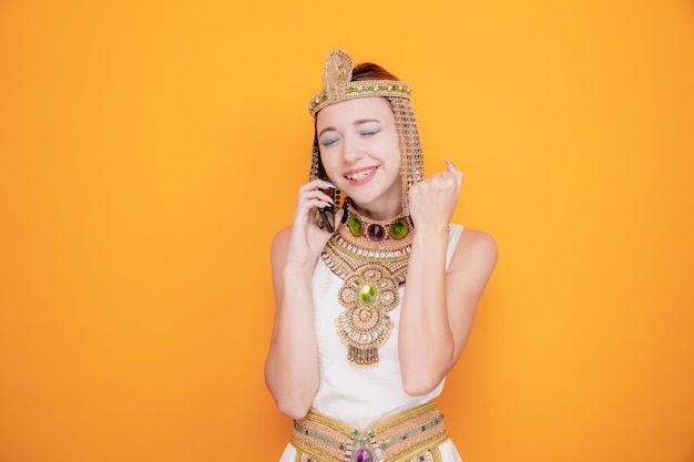 Beautiful woman like cleopatra in ancient egyptian costume happy and excited raising fist while talking on mobile phone on orange