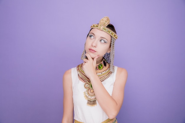 Free photo beautiful woman like cleopatra in ancient egyptian costume looking up puzzled with hand on her chin on purple