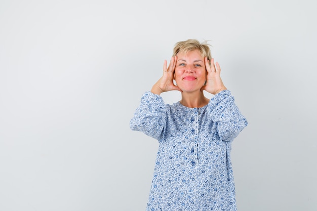 Beautiful woman in patterned blouse touching her cheeks with her hands and looking glad , front view. space for text