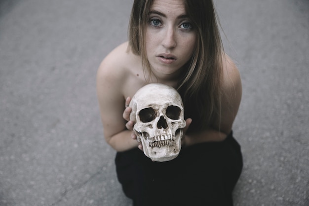 Free photo beautiful woman sitting on asphalt holding decorative skull and looking at camera