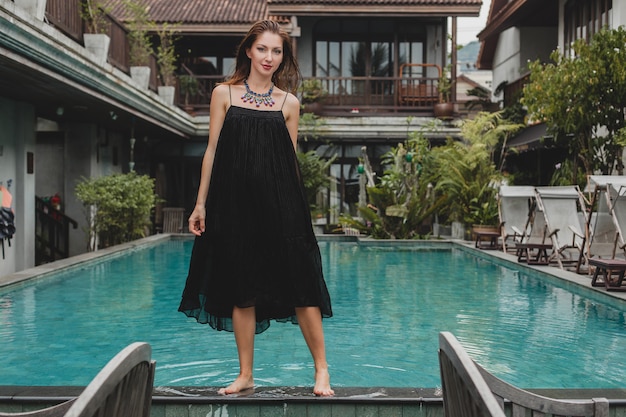 Free photo beautiful woman in stylish long black dress posing at tropical villa pool, elegant summer style, vacation, fashion trend, walking barefoot