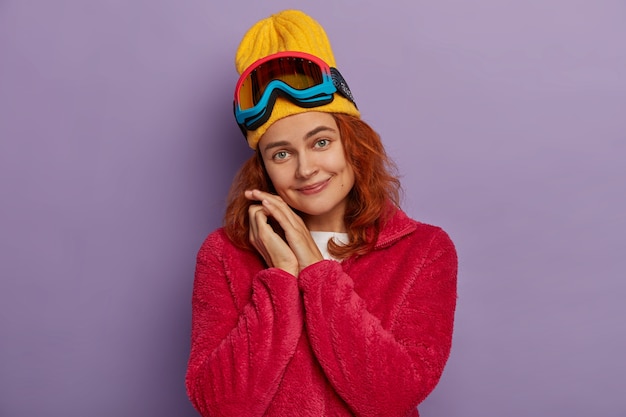 Free Photo beautiful woman tilts head, wears yellow hat and red jacket, has fun, uses snowboarding glasses looks gladfully at camera isolated on violet wall