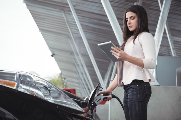 Beautiful woman using digital tablet while charging electric car