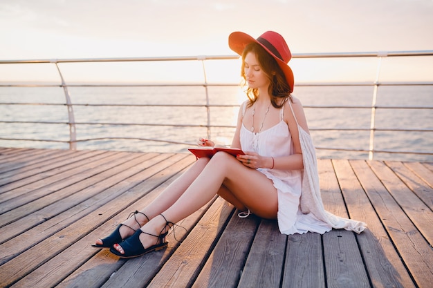 Free Photo beautiful woman in white dress sitting by the sea on sunrise thinking and making notes in diary book