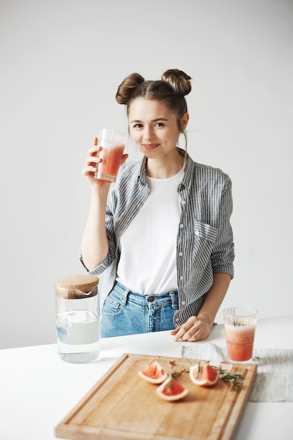 Free photo beautiful woman with buns smiling drinking grapefruit detox smoothie over white wall. healthy diet nutrition