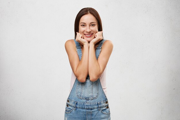 Beautiful woman with charming smile having joyful expression on her face, holding hands on her face, pleased with a compliment or nice present.