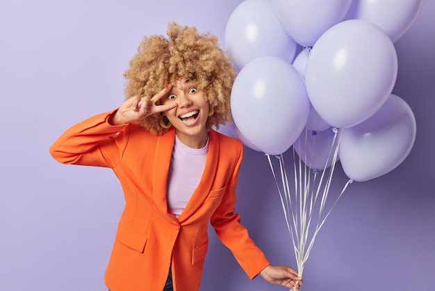 Beautiful woman with curly blonde hair makes peace gesture over eye dressed in fashionable orange jacket celebrates special occasion holds bunch of inflated balloons isolated over purple background