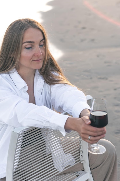 A beautiful woman with a glass of wine on the seashore sits on a chair