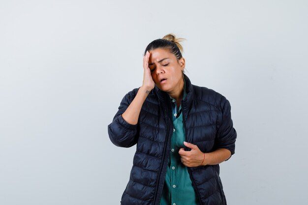 Beautiful woman with hand on temple, putting hand on jacket in green shirt, black jacket and looking harried , front view.