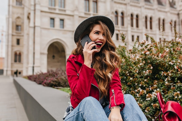 Beautiful woman with long wavy hairstyle talking on phone on architecture wall