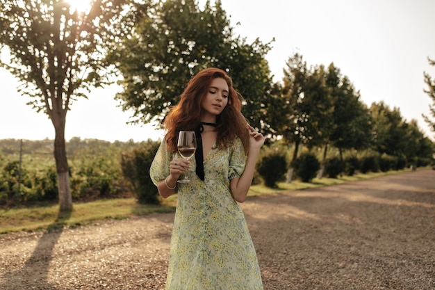 Free photo beautiful woman with red fluffy hairstyle and black bandage on neck in stylish summer dress holding glass with wine and hat outdoor