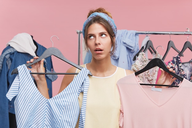 Free photo beautiful woman with tired expression holding two hangers with dresses choosing between two. discontent young female seller offering clothes in boutique, being exhausted of fastidious clients