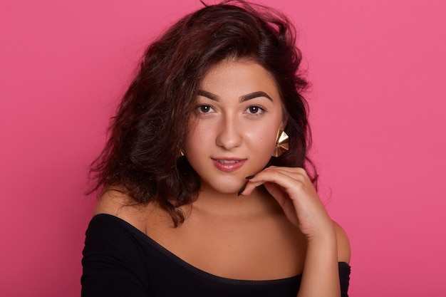 Beautiful woman with wavy brunette hair and clean skin with natural makeup posing with hands under chin against pink wall,  with confident expression.