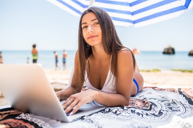 Beautiful woman working online on laptop while lying on beach under sun umbrella near sea. Happy smiling freelancer girl relaxing and using notebook for freelance internet work.