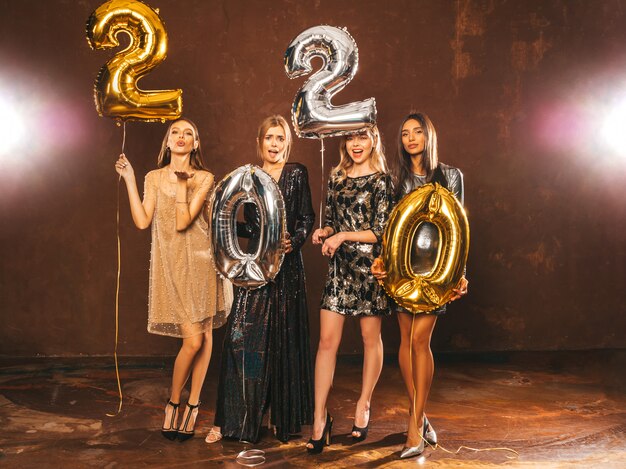 Beautiful Women Celebrating New Year. Happy Gorgeous Girls In Stylish Sexy Party Dresses Holding Gold and Silver 2020 Balloons, Having Fun At New Year's Eve Party. Holiday Celebration.Charming Models