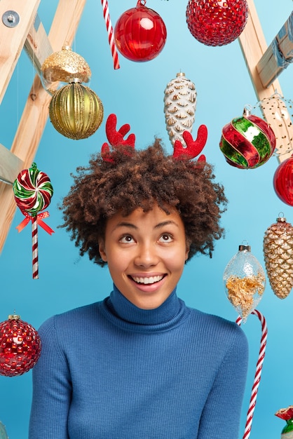 Beautiful young African American woman with curly hair looks above smiles gladfully dressed in casual poloneck surrounded by New Year toys 