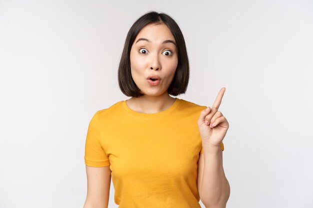 Beautiful young asian woman pointing finger up smiling and looking amused at camera showing advertisement announcement on top white background