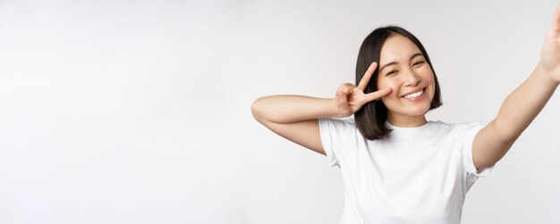 Beautiful young asian woman taking selfie posing with peace vsign smiling happy take photo posing against white background