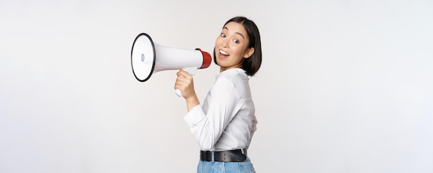 Beautiful young asian woman talking in megaphone screams in speakerphone and smiling making announcement shout out information standing over white background