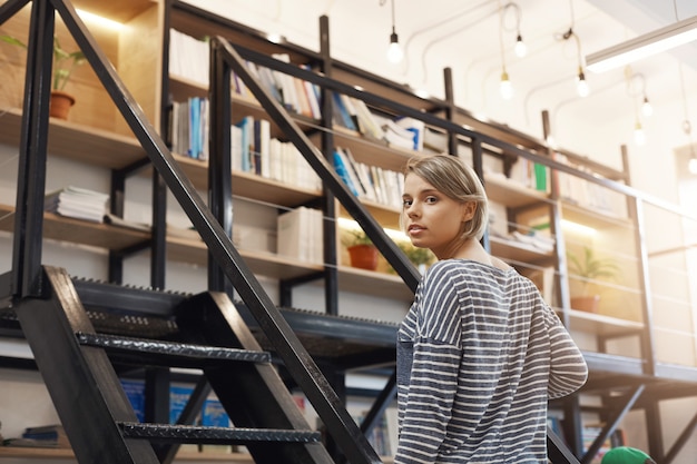 Free Photo beautiful young blonde student girl with short hair in casual striped shirt spending time in modern library after university, preparing for exams with friends. girl standing near stairs going to take