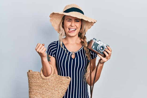 Beautiful young blonde woman holding vintage camera sticking tongue out happy with funny expression.