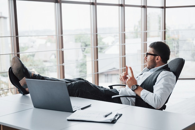 Beautiful young businessman with glasses holding his legs on the table looking at a laptop in the office