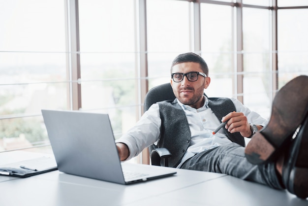 Beautiful young businessman with glasses holding his legs on the table looking at a laptop in the office