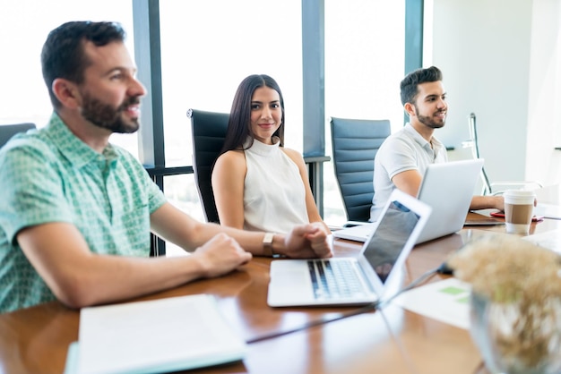 Free photo beautiful young businesswoman making eye contact while sitting amidst colleagues at desk during meeting in office