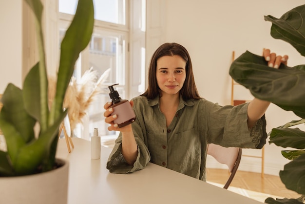 Free Photo beautiful young caucasian woman with jar of hair mask sitting at table indoors brunette in green shirt looks at camera beauty and care concept