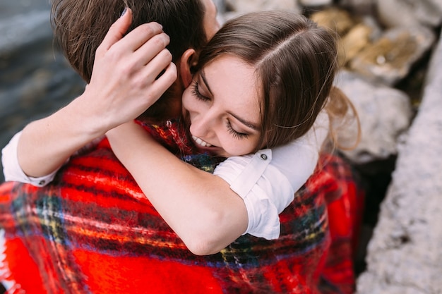 Free Photo beautiful young couple sitting on a stone embankment, wrapped in a blanket