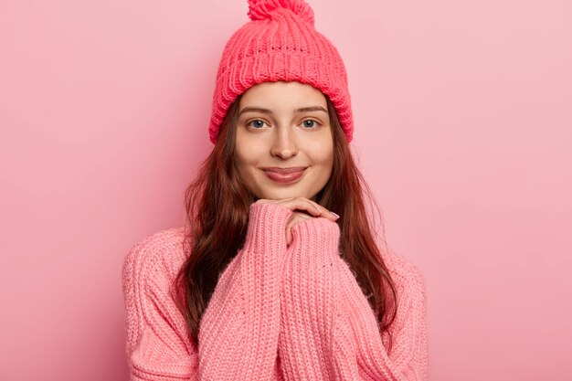 Beautiful young dark haired woman keeps hands uder chin, wears warm winter clothes, looks gladfully at camera has natural beauty with no makeup.